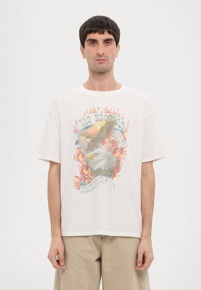 Camiseta de algodón blanca con un gráfico colorido de dos águilas rodeadas de llamas, con una pancarta que dice "DESDE 2003". Diseño de corte holgado.