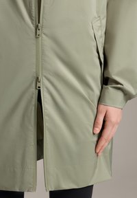 Jachetă waterproof verde deschis, cu fermoar frontal, buzunare laterale și manșete elastice. Textură netedă și design până la genunchi, cu croi drept.