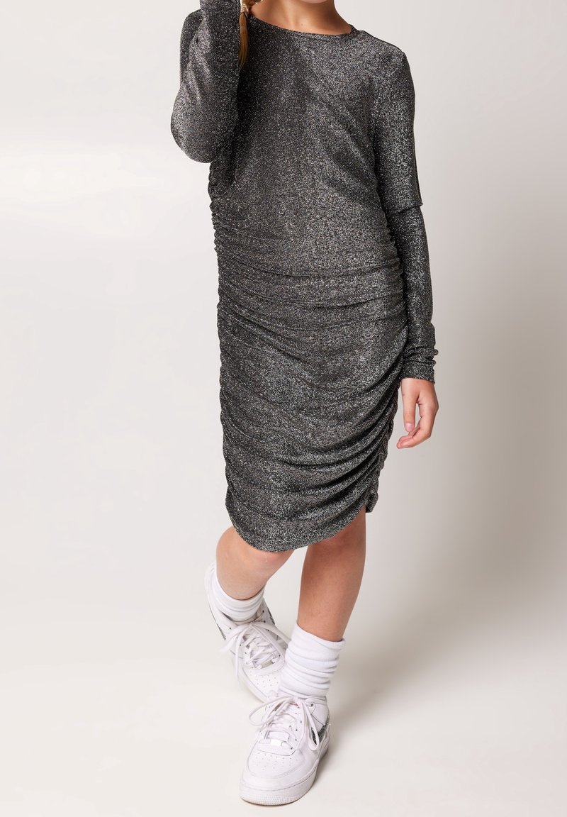 Fille portant une robe longue à manches longues, froncée, en tissu métallisé argenté, assortie de chaussettes blanches et de baskets blanches, debout avec une jambe légèrement avancée.