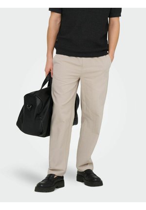 Osoba oblečená v béžových nohaviciach, čiernej košeli a čiernych topánkach, drží veľkú čiernu športovú tašku jednou rukou, druhá ruka v kapse.