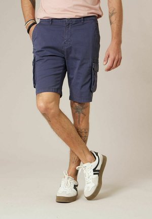 Homme portant un short cargo bleu marine, des baskets blanches avec des accents noirs, et des bracelets, debout les jambes croisées sur un fond uni.