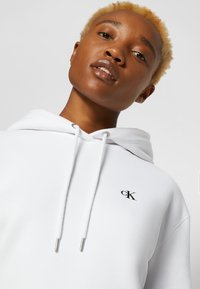 White hoodie made of soft fabric, featuring a drawstring hood and a small black "CK" logo on the chest. Smooth texture and modern design.