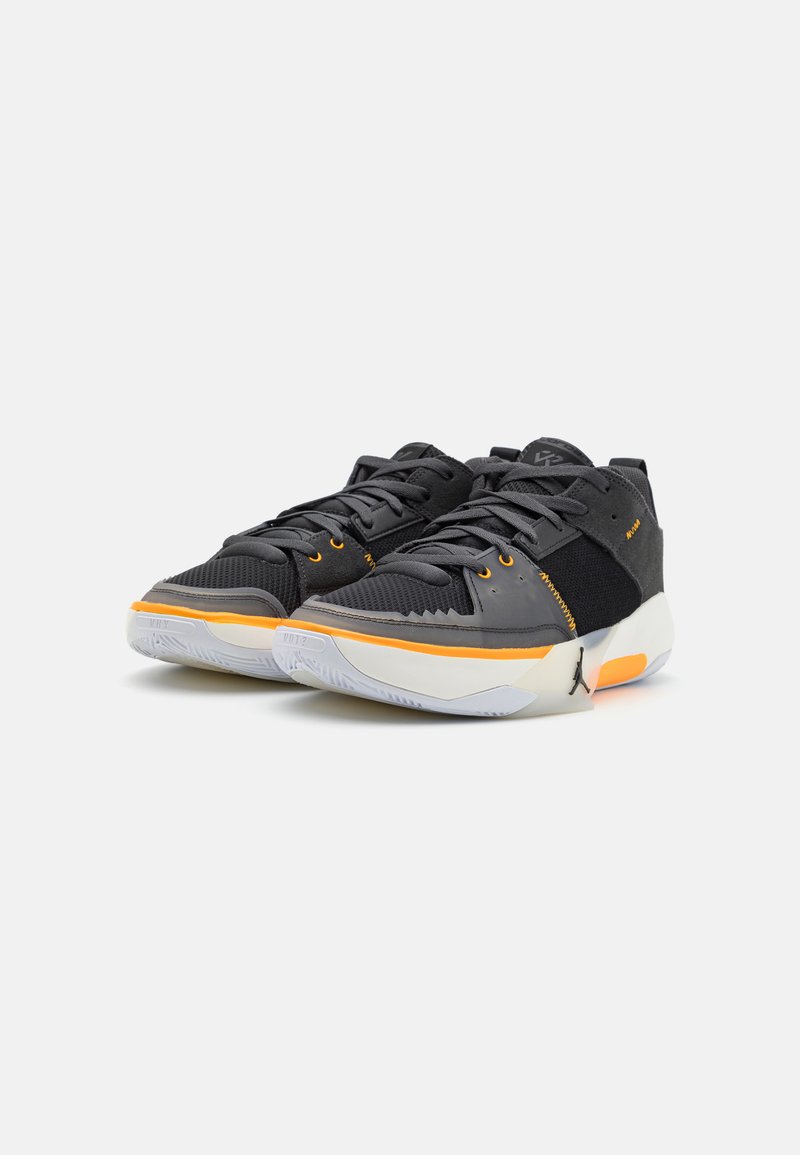 Zapatilla negra con materiales de malla y sintéticos, con acentos naranjas, diseño texturizado y una suela de goma blanca. Logotipo prominente en el costado.