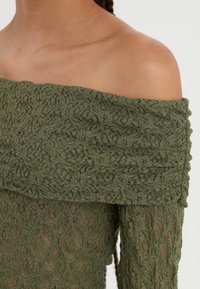 Ein schulterfreies Top in Olivgrün mit einer Spitzenstruktur und floralem Muster, aus weichem Strickmaterial und mit einem gerippten Saum.