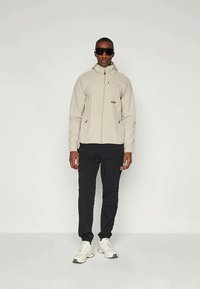 Vīrietis sagrāvisBeige kapucēts jaka, melni kargo bikses, baltas sporta kurpes un lielas melnas saulesbrilles, stāv uz viendabīgas fona.