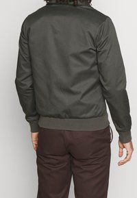 Giacca bomber verde scuro con una texture liscia, polsini e orlo a costine, dotata di un colletto classico e senza chiusure visibili o tasche.
