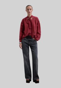 Mujer con el cabello recogido, vistiendo una blusa roja suelta con mangas acampanadas, jeans oscuros de pierna ancha, sandalias con plataforma y una pulsera de plata.