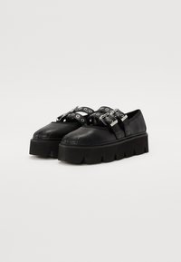 Zapatos de plataforma negros con suela gruesa, que presentan correas con herrajes plateados y recortes circulares, fabricados en cuero suave.