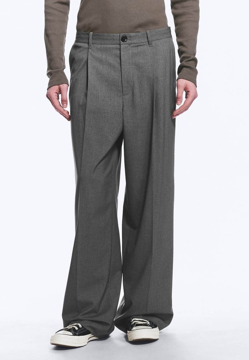 Homme portant un pantalon gris à jambes larges et des baskets noir et blanc, debout les bras détendus le long du corps.