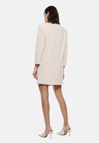 Cappotto beige su misura con maniche lunghe, taglio dritto e cuciture minimali. Indossato con tacchi chiari a punta.