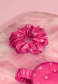 Różowa satynowa gumka do włosów z gromadzonymi teksturami i srebrnymi cyrkoniami, umieszczona na miękkim różowym tle.