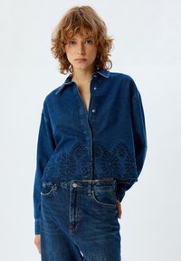 Camicia in denim blu con design cropped, caratterizzata da un intricata ricamo floreale all'orlo e chiusure con bottoni classici. Tessuto texturizzato.