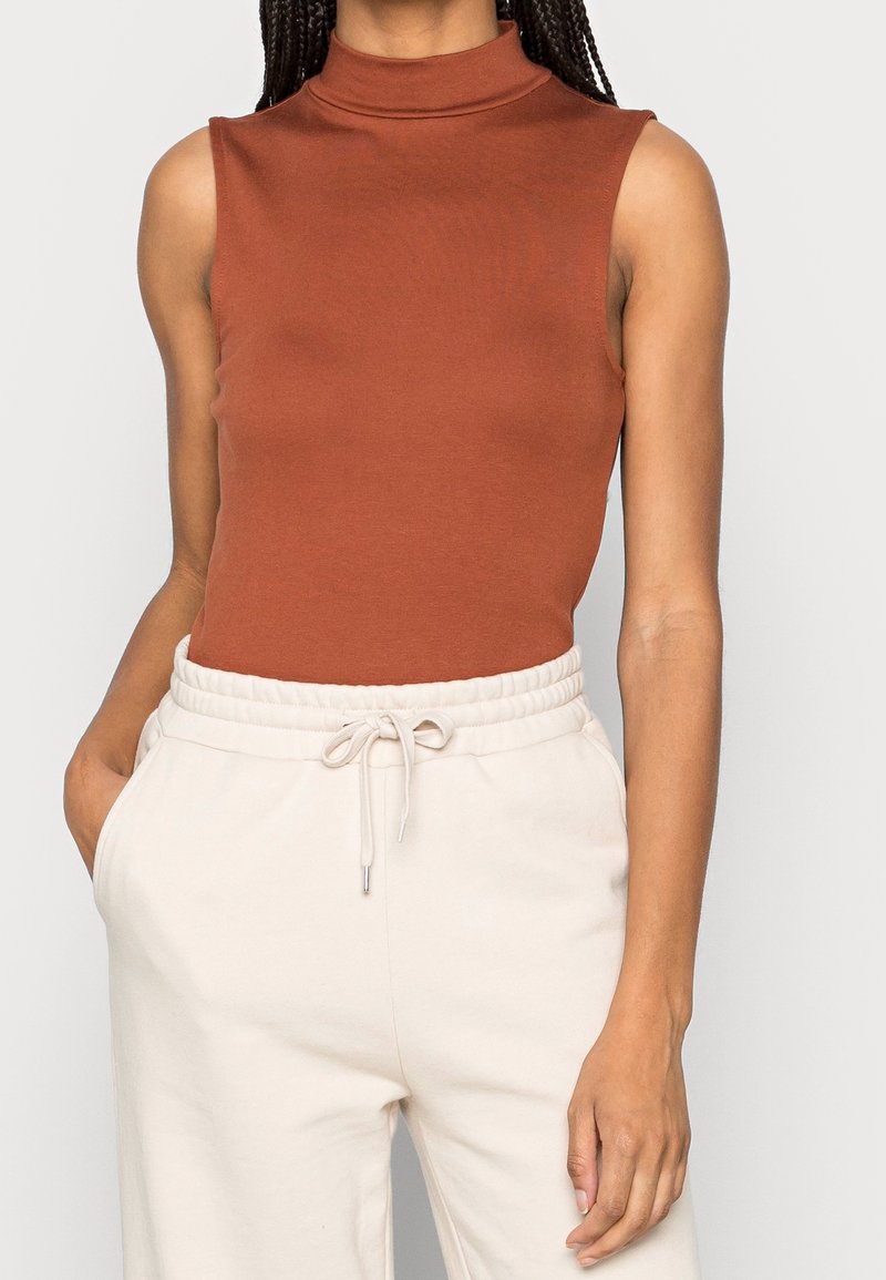 Haut sans manches marron à col montant, fabriqué en tissu lisse, associé à un pantalon de couleur claire à taille élastique avec poches latérales.