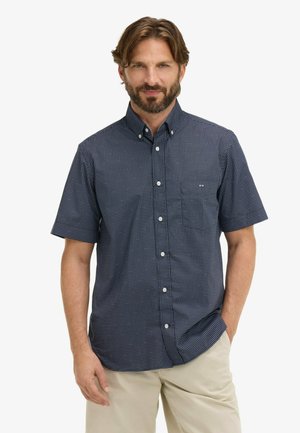 Homme barbu portant une chemise à manches courtes bleu marine à boutons avec un petit motif blanc et un pantalon beige, debout, main dans la poche.