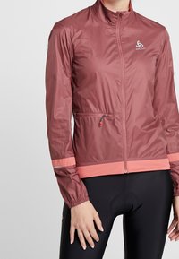 Letvægts, bordeauxfarvet vindjakke med høj krave, lynlåslukning, kontrasterende koralfarvede detaljer, lynlås-lomme og elastiske manchetter.