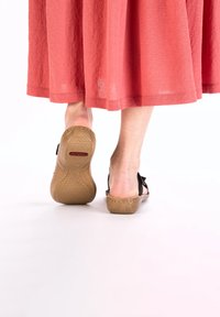 Des sandales noires à semelle texturée beige, dotées d'un design de sangle simple. L'arrière-plan présente une jupe rouge fluide.