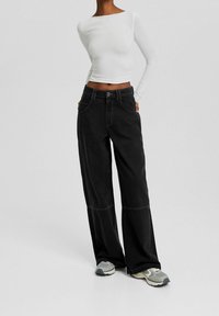 Wide-leg black jeans with pockets and contrasting stitching, paired with a fitted white long-sleeve top and gray sneakers.