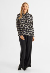 Blusa nera con un motivo geometrico crema, maniche lunghe e colletto. Abbinata a pantaloni neri larghi e scarpe a punta.