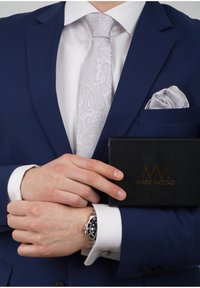 Marine-Anzug mit weißem Hemd, grauem Paisley-Krawatte, passendem grauen Einstecktuch, silberner Uhr, hält eine schwarze Box mit goldfarbigem Logo.