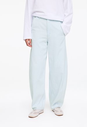 Persoon die lichtblauwe broek met wijde pijpen, een wit shirt met lange mouwen en witte sneakers draagt, staat met één hand in de zak.