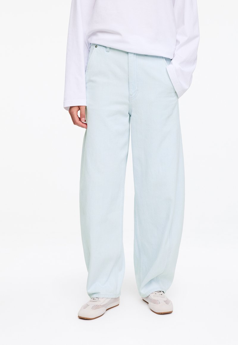 Personne portant un pantalon large bleu clair, une chemise blanche à manches longues et des baskets blanches, se tenant avec une main dans la poche.