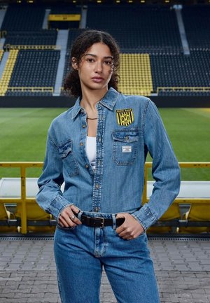 Mujer con chaqueta vaquera y vaqueros de pie frente a asientos vacíos de un estadio de fútbol con detalles en amarillo y negro.