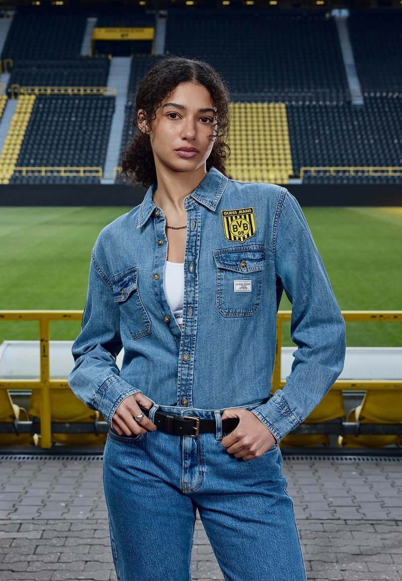Femme en veste en jean et jean, debout devant des sièges vides d'un stade de football avec des accents jaunes et noirs.