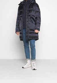 Abrigo acolchado azul marino con capucha, con diseño acolchado y cierre de cremallera, combinado con vaqueros azules y zapatillas blancas.