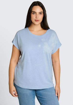 Femme aux longs cheveux sombres portant une chemise bleu clair à manches courtes avec une broderie florale sur le côté supérieur gauche et un jean bleu.