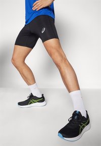 Shorts de sport noirs avec un design ajusté et un logo réfléchissant, associés à des chaussures noires avec des accents verts et des chaussettes blanches.