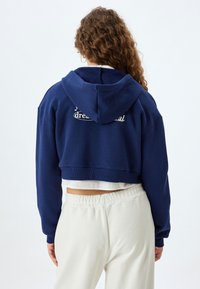 Sudadera recortada con capucha en color azul marino, fabricada en tejido suave, que presenta un corte relajado y un bordado en la parte posterior con el texto "sueña, hazlo". Pantalones blancos.