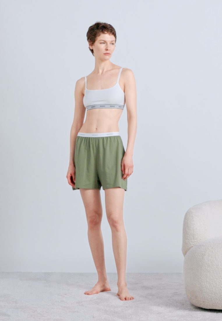Jeune personne debout pieds nus, portant un crop top blanc et un short vert, devant un fond uni avec une chaise blanche rembourrée à proximité.