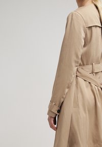 Trench beige con cintura ceñida, con un gran cuello, mangas largas y puños abotonados. Textura suave y ajuste entallado.