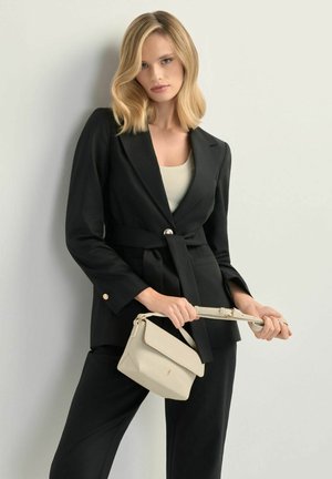 Kobieta w czarnym żakiecie z paskiem i spodniach, trzymająca beżową torebkę, stojąca na jasnoszarym tle.