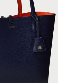 Μαρίνα δερμάτινη τσάντα tote σε σκούρο μπλε χρώμα με υφή pebble, πορτοκαλί εσωτερικό και χρυσή λεπτομέρεια λογότυπου. Διαθέτει δύο χερούλια και μια μικρή ετικέτα με charm.