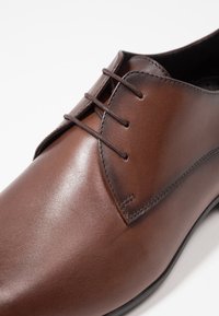 Bruine leren dresschoen met gesloten veters en een gladde gepolijste afwerking, getoond in close-up vanuit een hoek.