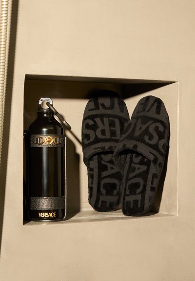Μαύρα παντόφλες Versace με ανάγλυφο λογότυπο, σε συνδυασμό με μια μαύρη μεταλλική φιάλη νερού με χρυσό λογότυπο και δερμάτινες λεπτομέρειες.