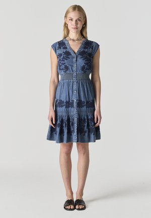 Mujer de pie, mirando hacia adelante, vestida con un vestido azul sin mangas abotonado con patrones florales oscuros y sandalias negras tipo slide.