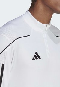 Närbild av en person som bär en vit atletisk zip-up-tröja med svart Adidas-logotyp och svarta sidostrekar på ärmarna.