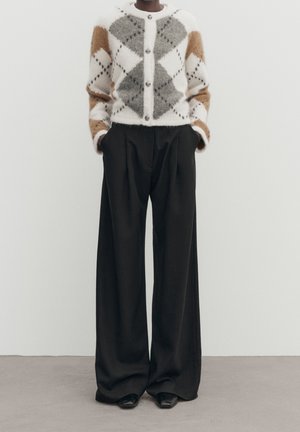 Personne portant un cardigan à motif losange blanc, gris et marron et un pantalon large noir, debout devant un fond uni.