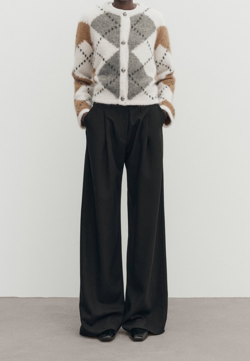 Personne portant un cardigan à motif losange blanc, gris et marron et un pantalon large noir, debout devant un fond uni.