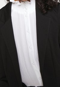 Valkoinen pusero, jossa on frillikaulus ja kangaspäällysteiset napit, käytetty mustan bleiserin alla, näyttäen ylävartalon ja osittain kiharat hiukset.