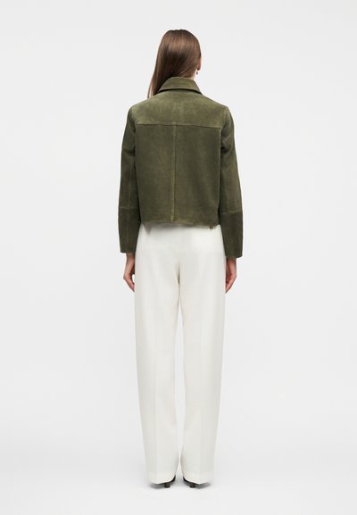 Γυναίκα με πλάτη προς τα πίσω, φοράει κομμένο χρυσαφί-πράσινο σακάκι και λευκό, ψηλόμεσο, φαρδύ παντελόνι σε μονόχρωμο φόντο.