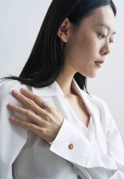 Bague en argent avec des détails complexes sur une main reposant sur une chemise blanche. La chemise présente un bouton doré au poignet. Éclairage doux.