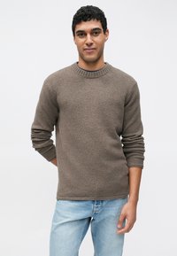 Ruskea teksturoitu neulepusero pyöreällä pääntiellä ja resorireunuksilla, käytetty vaaleansinisissä farkuissa. Malli seisoo käsien osittain selän takana.