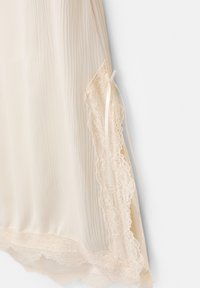 Rochie slip bej deschis din material plisat, cu dantelă pe margini și o fundă din satin pe lateral. Textură moale și formă fluidă.