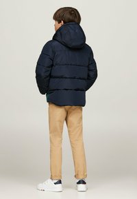 Giacca puffer blu navy con cappuccio, caratterizzata da un design trapuntato e chiusura con zip. Indossata con pantaloni beige e sneakers bianche.