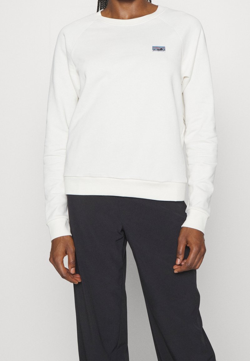 Personne portant un sweat-shirt blanc à manches longues avec un petit logo et un pantalon noir sur un fond uni.
