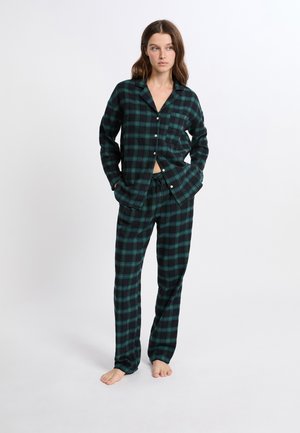 Femme debout pieds nus portant un haut de pyjama à carreaux noir et vert avec des boutons et un pantalon assorti, les mains dans les poches.