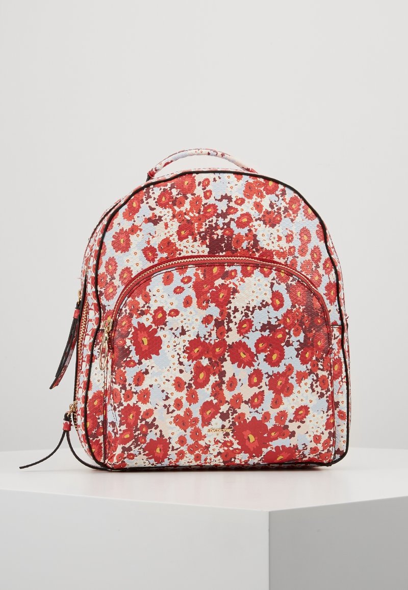 Petit sac à dos avec motif floral rouge et orange sur fond blanc, poche avant zippée et poignée supérieure, posé sur une surface blanche.